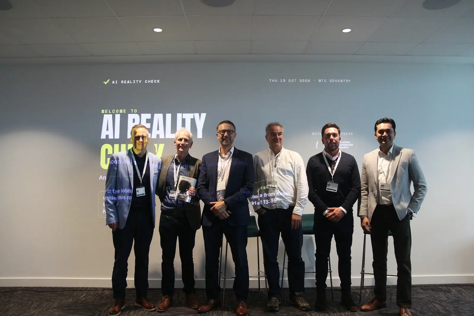 Speakers and host lined up in front of the AI Reality Check wall at the MTC